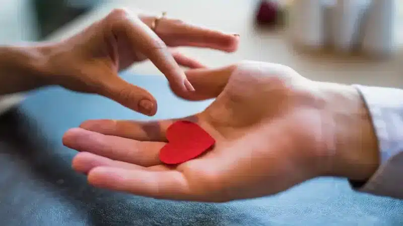 A hand extends towards another, which holds a small red felt heart, symbolizing love and connection between two people A hand extends towards another, which holds a small red felt heart, symbolizing love and connection between two people